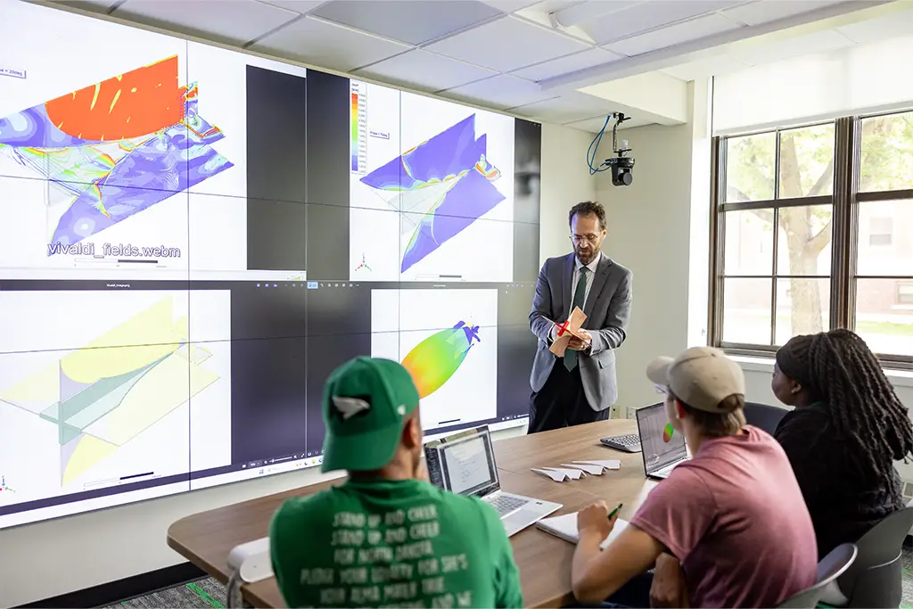 In a high-tech lab, an engineering professor discusses advanced research with Ph.D. candidates who are analyzing complex 3D models on a large video wall.