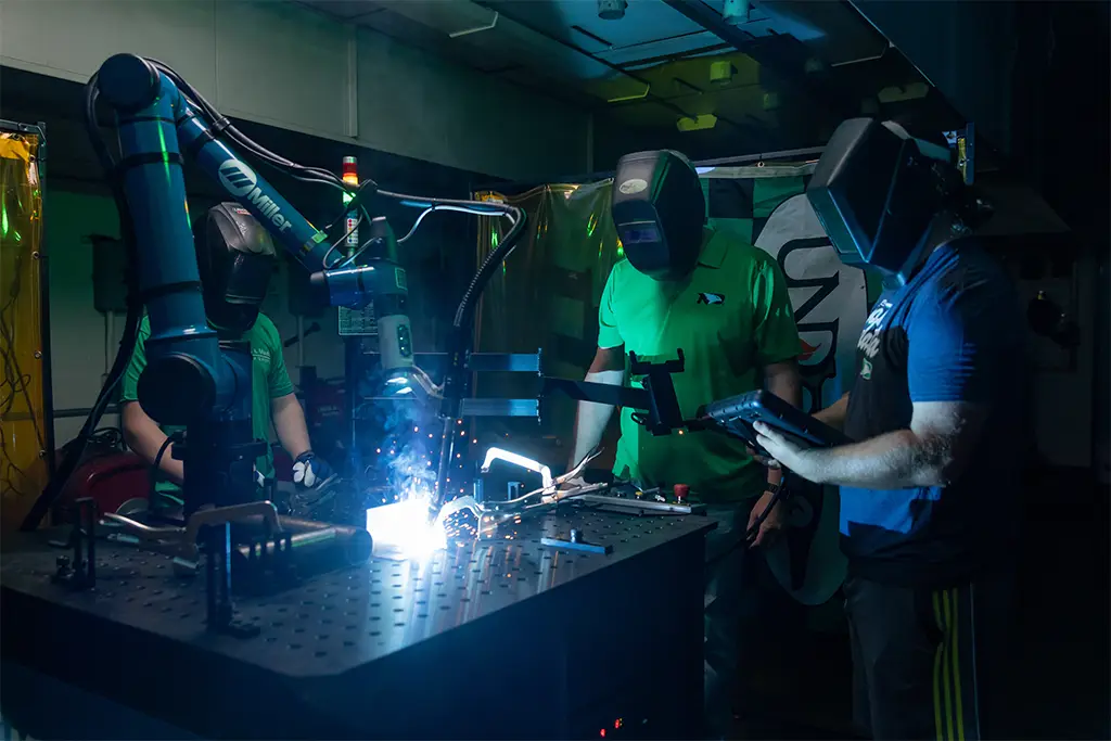 In a high-tech lab, engineering graduate students wearing protective helmets collaborate to operate a robotic welding arm, demonstrating the advanced, hands-on specialization acquired in a master's degree program.