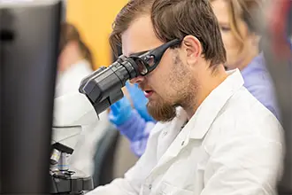 A medical laboratory science student in a white coat looks into a microscope in a busy lab.