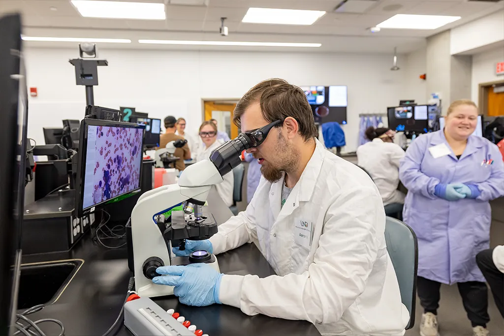A medical laboratory science student examines a blood smear at a microscope, the cells shown on a monitor.