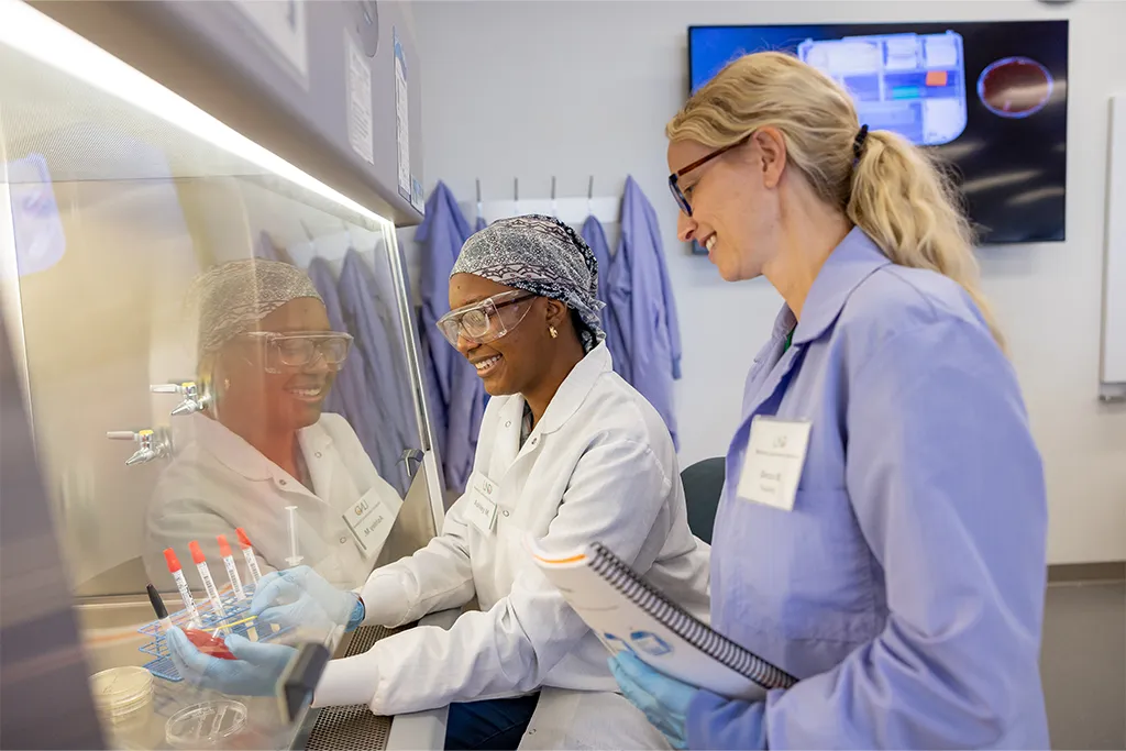 Two students in lab coats and goggles smile while handling blood sample tubes at a fume hood.