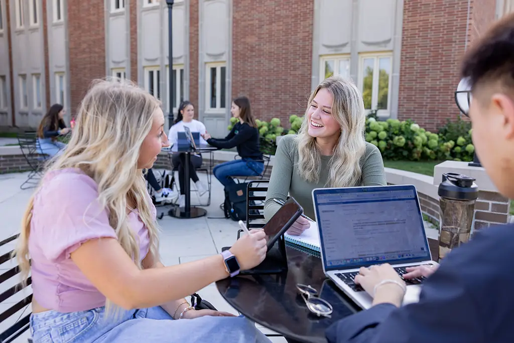 Three students sitting outside a university building with laptops and a tablet, checking online if a program meets ABET accreditation standards, while other students study in the background.