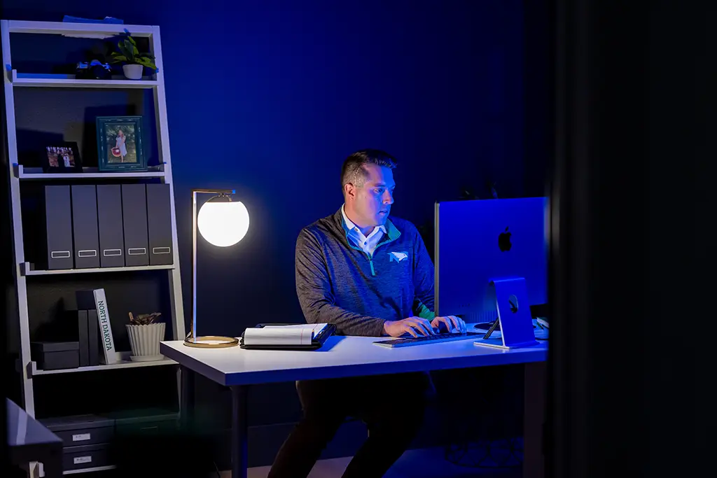 Man sitting alone in a dimly lit office at night, illuminated by a desk lamp and computer screen, reviewing reports and data that represent accountability and oversight in AI governance