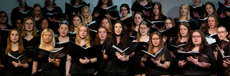 A women's choir in black formal attire sings from black folders during a concert performance.
