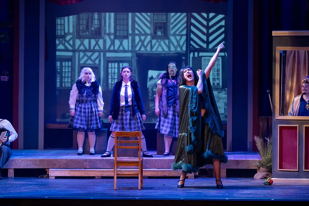 Costumed performers in plaid school uniforms watch a singer in a cape belt out a number on a stage set.