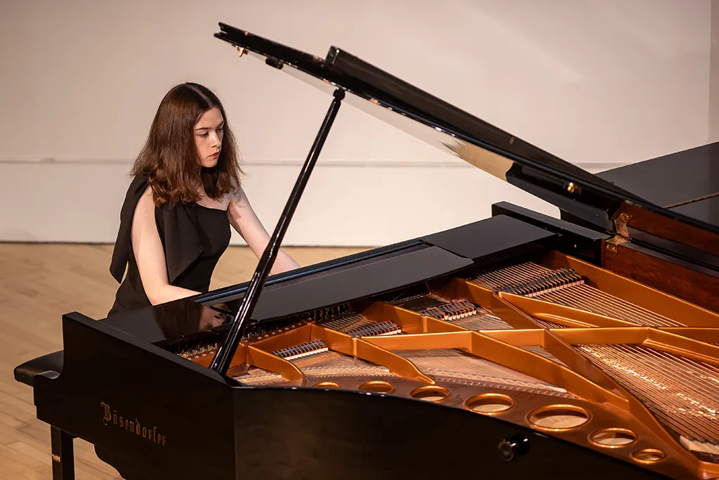 A student in a black gown performs at a Bösendorfer grand piano on a recital stage.