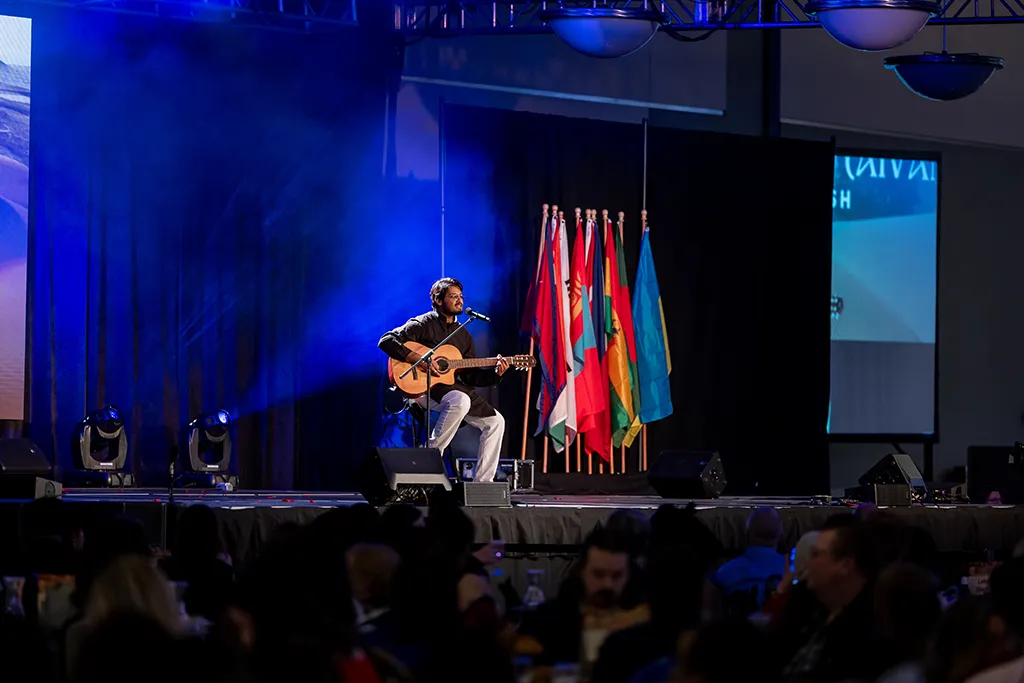 A musician plays acoustic guitar and sings onstage beside a row of flags before a seated audience.