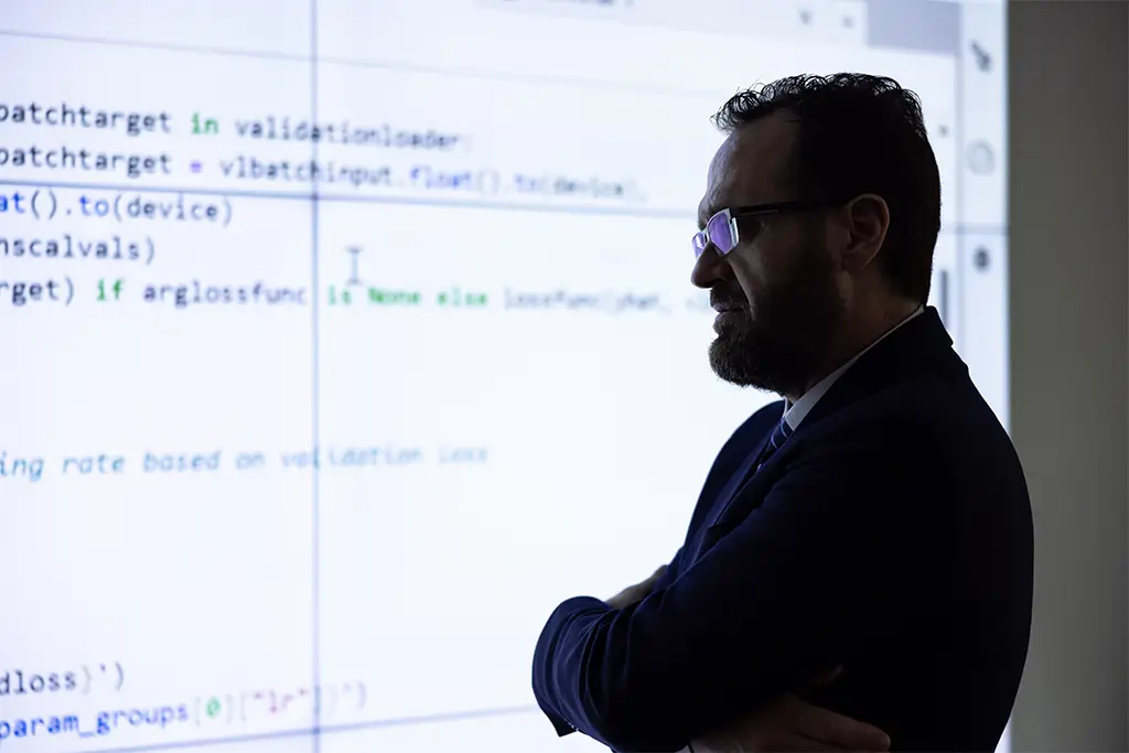 Man standing with folded arms in front of a large screen displaying lines of AI programming code, symbolizing thoughtful evaluation and the integration of ethics into every stage of AI system development.