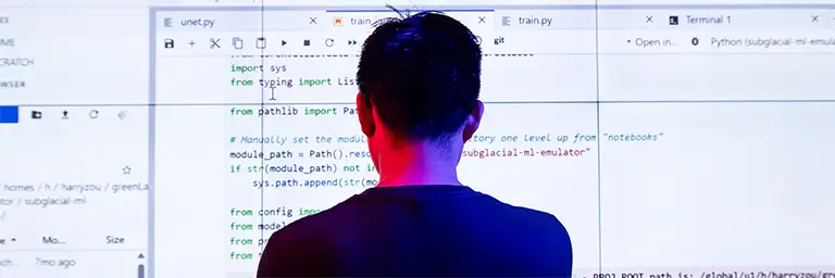 Student standing in front of a large screen displaying AI model code, symbolizing the link between artificial intelligence, technology, and ethical responsibility.