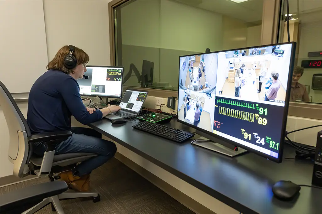 A technician wearing a headset sits at a desk in a monitoring room, observing multiple computer screens displaying live video feeds and vital sign data from a medical simulation or clinical setting