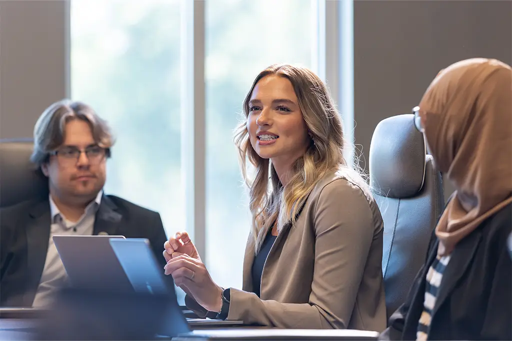 Professionals sit around a conference table during a business meeting. A woman speaks confidently while colleagues listen, reflecting career advancement and leadership in engineering.