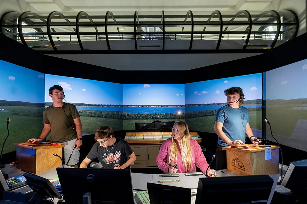 Four air traffic control students operate the tower simulator with panoramic runway screens behind them.