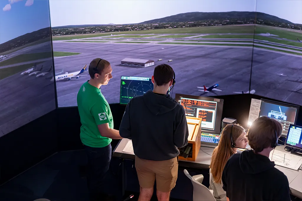 Air traffic control students in headsets monitor traffic at consoles facing the tower simulator screens.