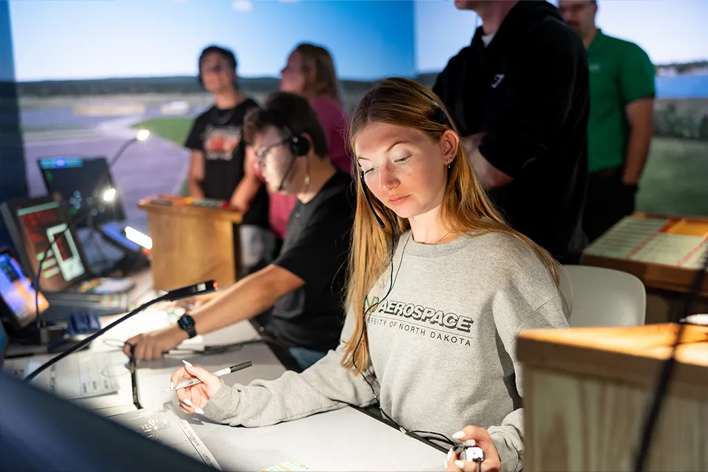 A student in a UND Aerospace sweatshirt and headset marks flight strips in the tower simulator.