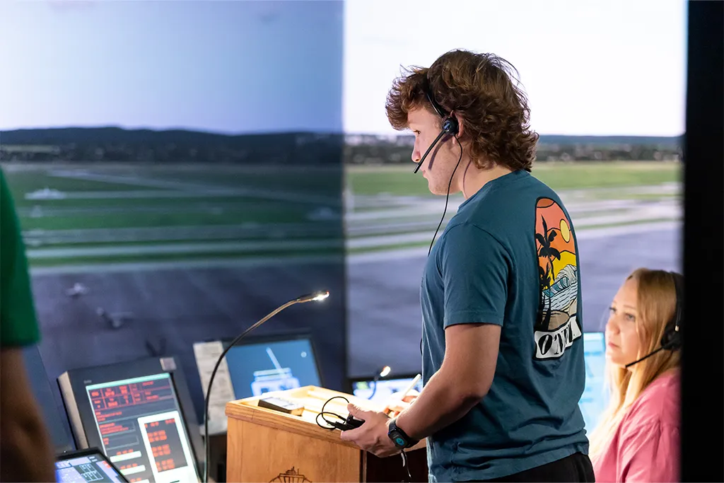 An air traffic control student in a headset works the control panel facing the tower simulator screens.