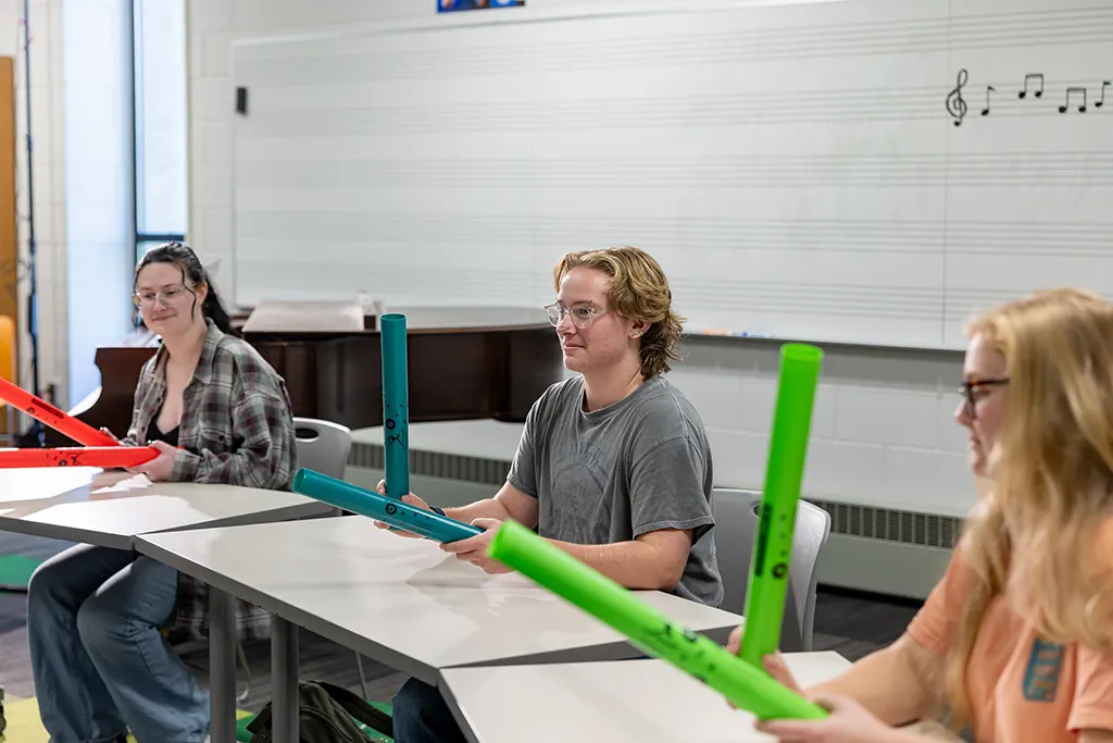 Three music therapy students hold colorful boomwhacker tubes at desks in a music classroom.