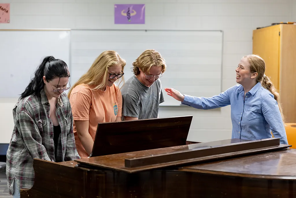 An instructor laughs with three music therapy students gathered around an upright piano.