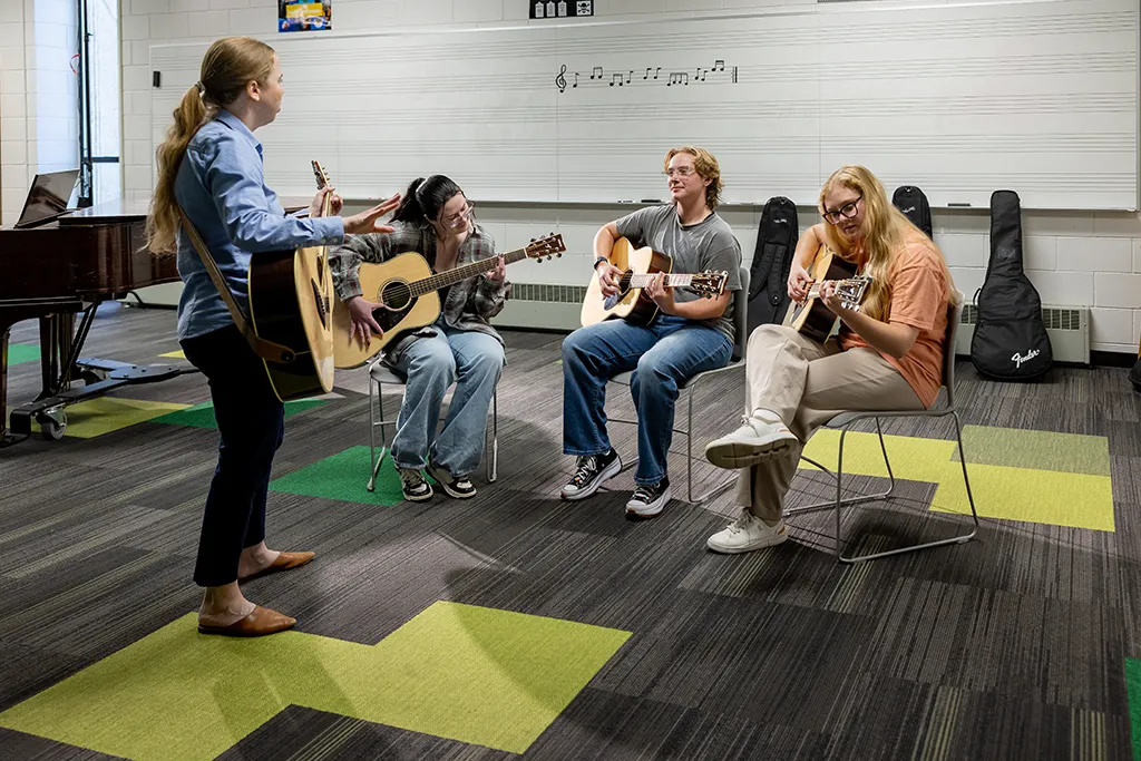  An instructor with a guitar leads three students playing acoustic guitars in a music classroom.