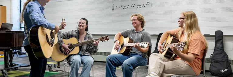 Three students and an instructor play acoustic guitars together in a music classroom.