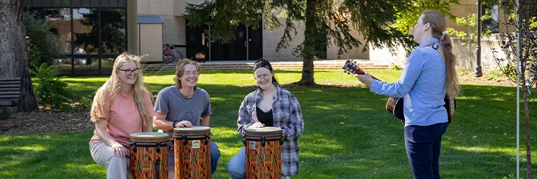 Three students play djembe drums on a campus lawn while an instructor accompanies them on guitar.