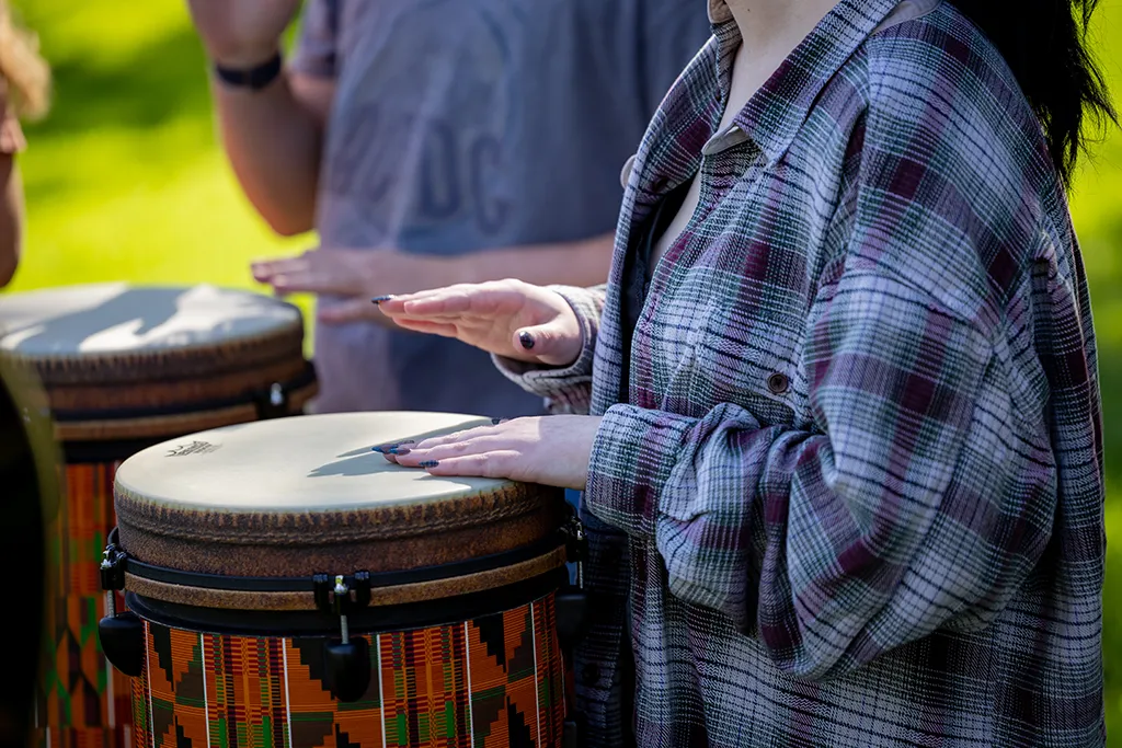 Close-up of a student's hands playing a kente-patterned djembe drum outdoors.