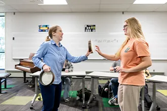 An instructor hands a frame drum to a student in a music classroom while others sit at tables.
