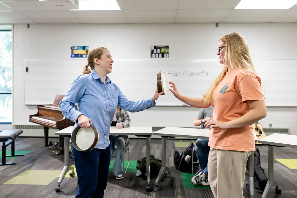 Music therapy instructor and student practice rhythm with hand drums in a classroom.