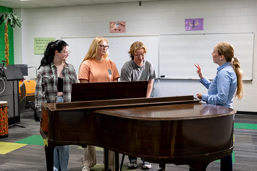 An instructor gestures while teaching three music therapy students gathered around a grand piano.