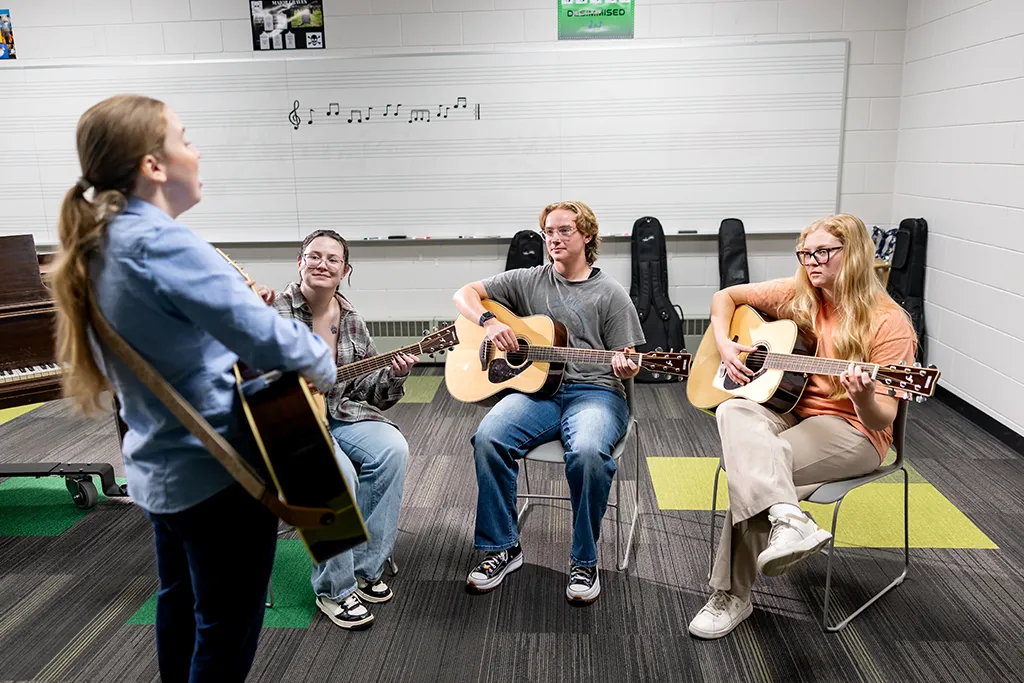 An instructor with a guitar leads three music therapy students playing acoustic guitars in a classroom.