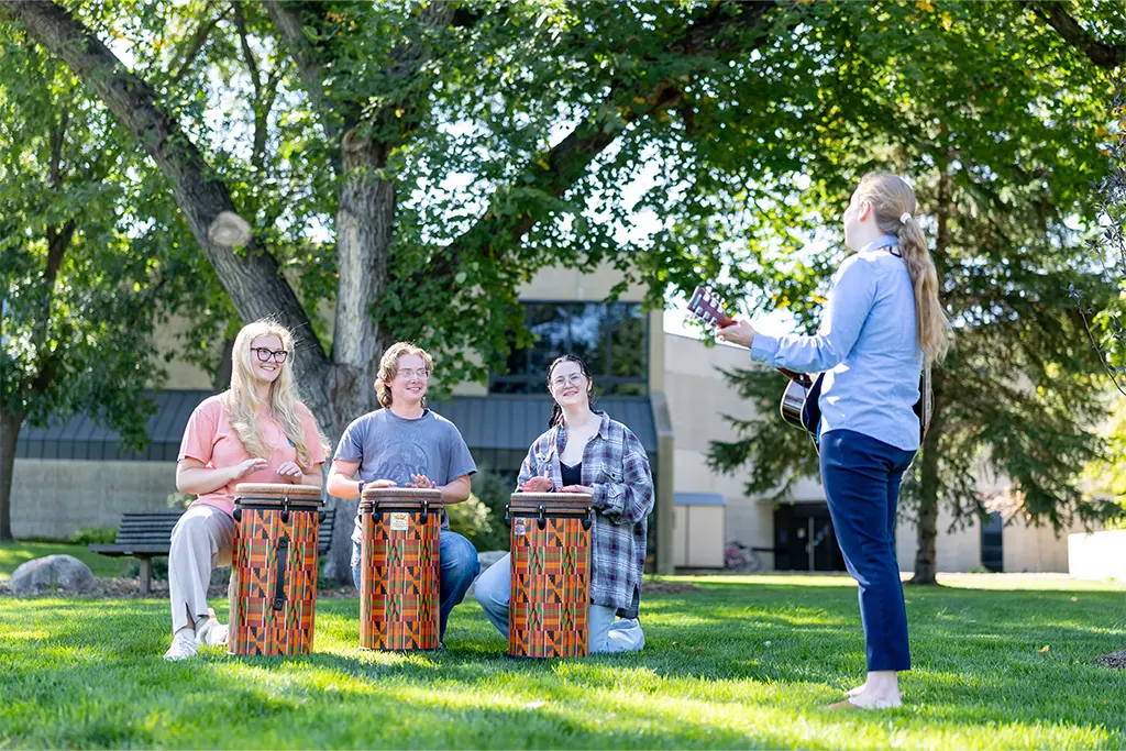 Three students play djembe drums outdoors on a campus lawn while an instructor accompanies them on acoustic guitar