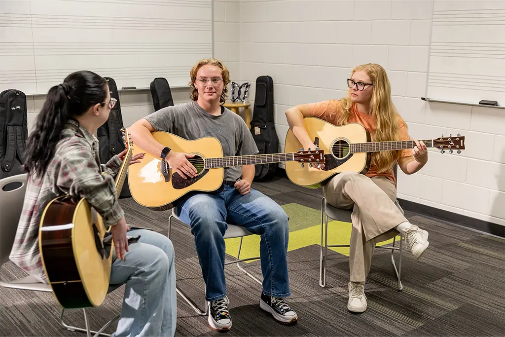 Three music therapy students practice acoustic guitar together in a rehearsal room with instrument cases lining the wall.