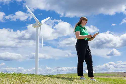 student standing with a clipboard with a wind turbine in the background