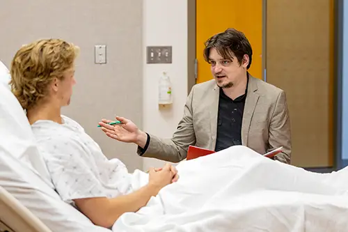 Chase Garber holding a folder sitting next to patient in bed talking