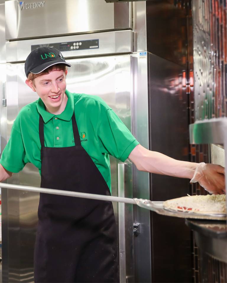 dining student employee placing a pizza into a pizza oven