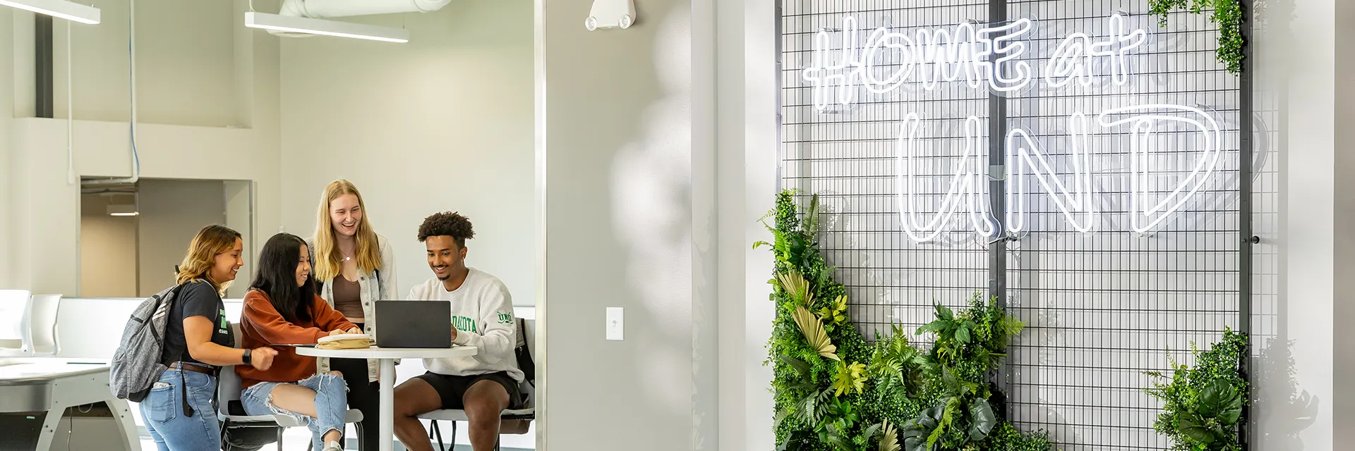 Students gather at a computer in a large study space with a Home at UND neon sign in the foreground