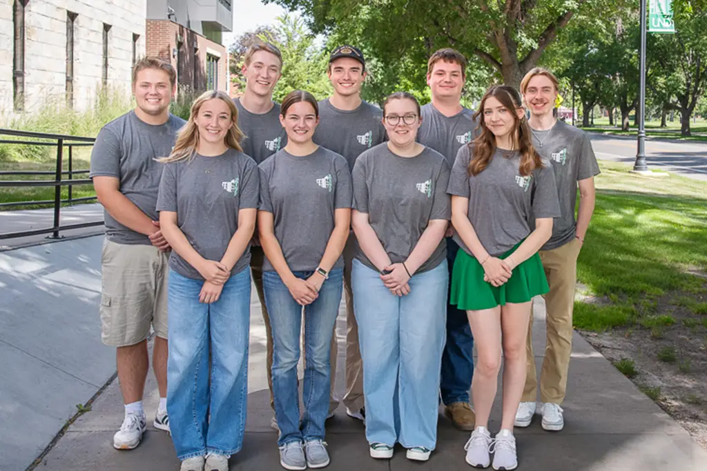 Group of young adults in matching uniform t-shirts pose on a sidewalk outside on a sunny day.